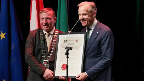 Reuters Two men holding a large framed script. The man on the right is wearing a navy suit and green tie. The man on the left is wearing a mayoral chain. 