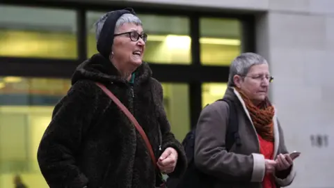 Alyson Lee (right) and Di Bligh leaving the environs of Westminster Magistrates' Court. One looks cheerful and is wearing a furry jacket and an orange cross-body bag, while the other seems more serious and wears a grey coat with an orange jumper and russet scarf underneath. Both women have short grey hair.