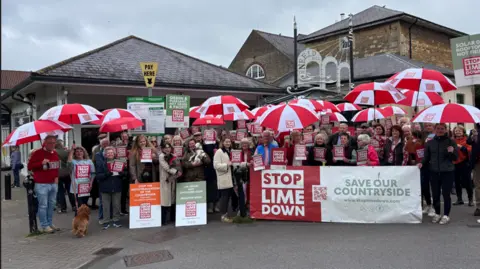 A large group of people stand with banners and umbrellas which say 'Stop Lime Down' with other slogans such as 'Save Our Countryside'