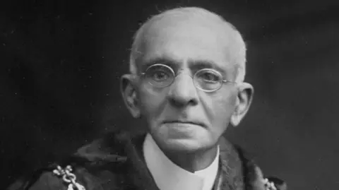 A black and white photo of Kavas Jamas Badshah. He is an elderly man wearing small round frame glasses and looking at the camera.