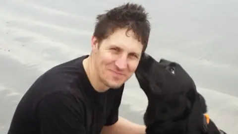 Jodie Smith A man is on a beach with a black dog licking the side of his face. The man is wearing a black T shirt.