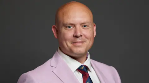 A bald man smiles at the camera. He is wearing a white shirt, blue and red tie, and a pink suit jacket.