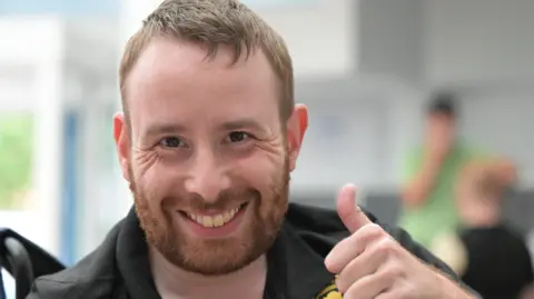 A man with short light brown hair is giving a thumbs-up gesture while seated indoors. He is wearing a black polo shirt with gold embroidered logos, including one circular badge on the chest. The background is softly blurred, showing another person in green.