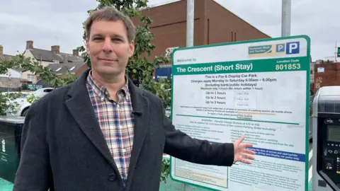 Councillor Wilkins is stood looking at the camera in a car park, gesturing at the car parking board behind him. He's wearing a checed shirt and grey overcoat.