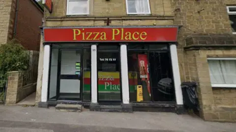 Front view of a takeaway restaurant in a stone-built terrace building. A large red sign above the shopfront reads “Pizza Place” in yellow lettering. The premises feature black-framed windows and doors separated by white columns, with additional text on the glass including a telephone number. A colourful panel inside the window also displays the business name. A yellow floor warning sign is visible near the entrance, and two wheelie bins stand beside the building on the right. The shop sits directly on a pavement along a street lined with older stone properties.