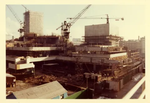Sir Robert McAlpine Cranes and scaffolding above the part-built concrete frame of the National Theatre on the South Bank in 1970. There are lots of levels and sections. Behind the construction site are other buildings. 