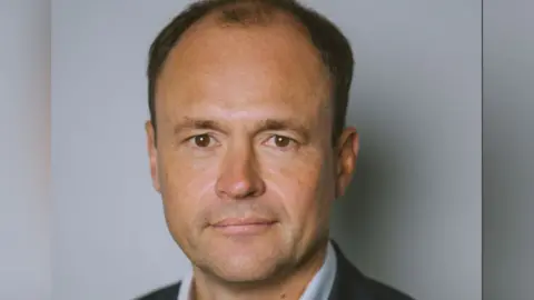 SpaMedica A headshot of David Hare who is stood in front of a grey background. He has brown eyes and is clean shaven. He appears to be wearing a suit and shirt, but only the top of the collar is visible. 