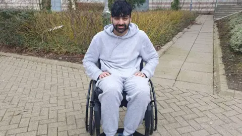 A young man with dark hair and facial hair sits, smiling at the camera. He is dressed in a grey hoodie and sweatpants. He is outside.