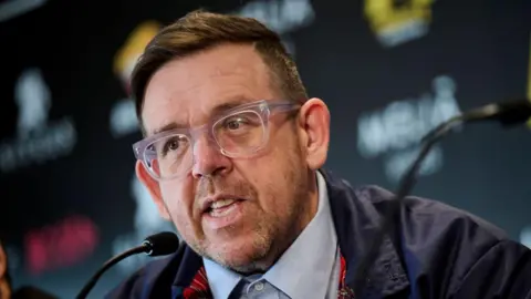 Nick Frost wearing glasses in front of a black background. He is talking into a small microphone.