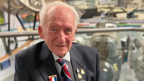 Jeff Brown is wearing a dark suit and blue/red tie. He has his operational medals and pin badges on his jacket. 