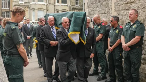 Paramedics watch on a a coffin is carried to a church