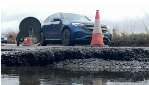 A view from the pothole, which is deep, and filled with water, looking up towards the road. There are traffic cones and signs on the road, and a car drives past.