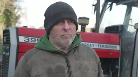 A man, with grey stubble, wearing a dark grey hat, green hoodie and green jacket. He is standing in front of a red tractor.