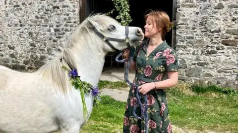 A white pony on the left looks up to Donna Oldbury-Thomas who is looking at the pony wearing a green dress with pink flowers and brown boots.