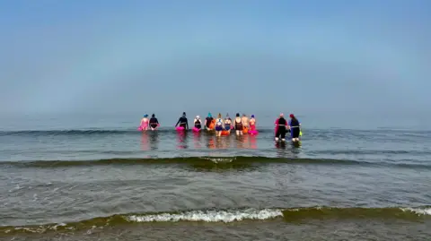 ELENA REID A group of 12 people in the water at Nairn Beach. Most are wearing swimsuits in various colours but four are wearing full body wetsuits in dark colours. All are wearing swimming caps. The water is calm and blue coloured, like the sky. A wave with white foam is rolling in in the foreground.