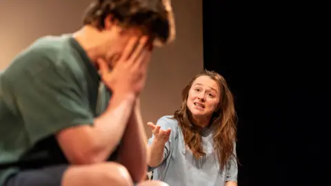 Johan Persson Sheehy wears a light blue baggy t-shirt. She looks angry, and is pointing to Aramayo, who sits with his head in his hands wearing a green t-shirt. 