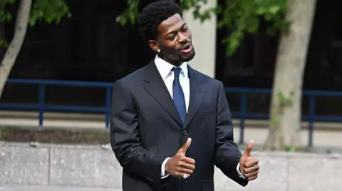 Hill flashed thumbs up to reporters after the hearing. Nas X wearing a suit and with his thumbs up and smiling