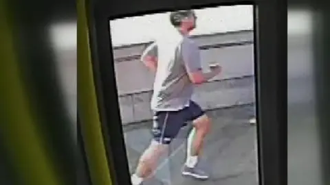 CCTV still image from the inside of a bus showing a man in a grey T shirt and shorts running. 