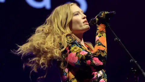 Freya Ridings with a mic in her hand. She has long blonde hair and is wearing a top with a floral pattern.