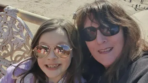 Family Sandra with her daughter Beatrice. They are taking a selfie by the seaside. There is a beach behind them. They are both wearing sunglasses. 