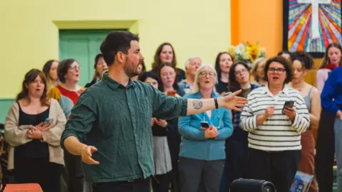 Whale Wise A man in a green shirt gesturing. Several women are stood in rows behind him and appear to be singing. They are in a room with brightly coloured walls.