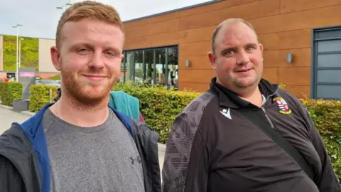 Two men look at the camera, smiling. One has short ginger hair and is wearing a grey t-shirt with a black jacket unzipped down the middle. He is standing next to a man with a balding head and wearing a black sport's top with a bag across his chest.