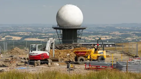 Julian Cartwright A huge golf-ball like radar on top of a large platform with a fence around its perimeter. The dome is white and has hexagonal shapes on its outside. The tower is surrounded by metal fencing and on the ground below it are mounds of earth There is a small yellow earth mover with a person sitting on it with a red helmet. There is also a small red and white digger