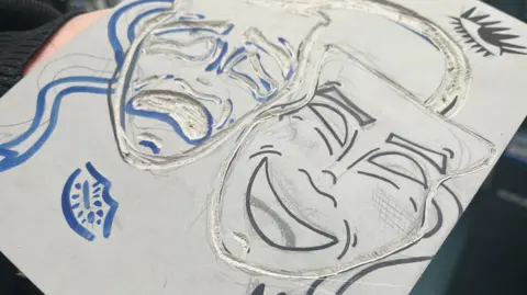 Jon Wright/BBC Two masks, one smiling and one frowning, are carved into a grey lino square