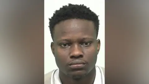 Mugshot if Ishiekwenne. He has very faint black stubble on his chin and upper lip and short thick black hair.