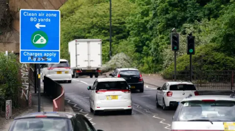 Two lanes of cars next to a large clean air zone sign.