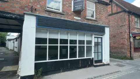A vacant shop unit with black strips above and below the plain windows. Above the shop is a red brick floor with a for sale sign attached.