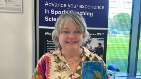 BBC / Jack Hadaway-Weller A person is standing indoors in front of a display poster and a window overlooking a sports field. The poster behind them reads “Advance your experience in Sports Coaching Science MSc?” with additional smaller text below. The person is wearing a patterned shirt and smiling toward the camera, with bright natural light coming through the window.