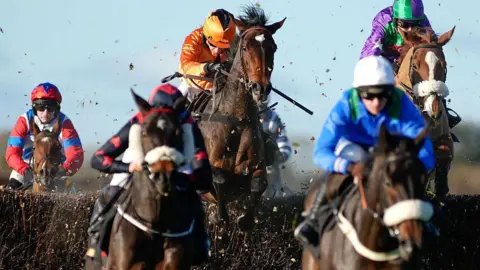 Getty Images Jockeys and their horses jump over a hurdle, with debris flying around. The jockeys are wearing colourful uniform.