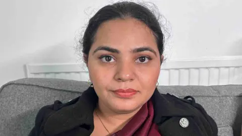 Kate Bradbrook/BBC Close-up of a woman sitting on a grey sofa indoors with a radiator behind.
