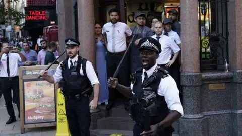 Aaron Chown Two police officers stand at a doorway of a McDonalds holding batons in front of staff standing on the steps