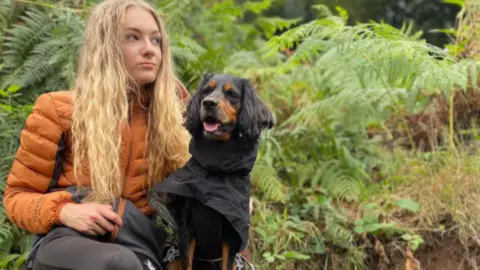 Family Handout Lily-May sits on a bench in woodland with a brown and black dog. She has an orange puffa jacket on and long curly blonde hair and leggings on.