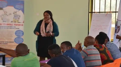 Supplied Cynthia Waggah giving a presentation in Kenya as part of her former government job. She is wearing a black dress with a light scarf wrapped round her neck. Several men are sitting in a row listening to her speak.