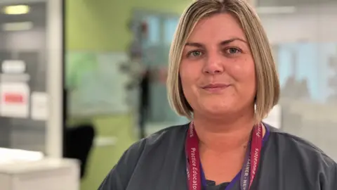 BBC Louise Lightfoot has blonde bobbed hair and its wearing grey scrubs with red and violent lanyards identifying her roles. A blurred shot of a ward is in the background.