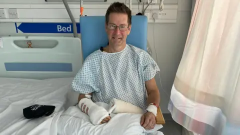 A man is sitting in a blue hospital chair next to a bed with a blue and white long top on. His right arm is bandaged and his left has a bandage around his wrist. He is wearing rectangular glasses and has short brown hair which is slightly spiked up.