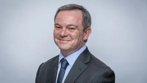 A head and shoulders photograph of Ian Haythornthwaite wearing a grey suit, light blue shirt and blue-spotted tie. He is smiling while looking slightly side on at the camera. 