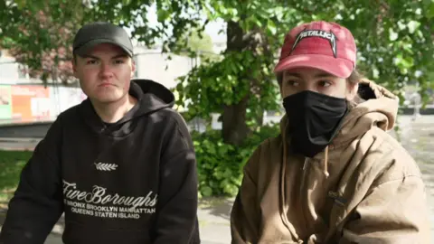 Two young men sitting down, one wearing and black hoodie and black cap, and the other wearing a red cap, with the bottom half of his face covered by a black snood