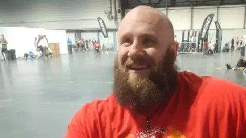 Matt Smyth smiles, looking just off camera, after winning the competition. He is in a mostly empty warehouse gymnasium. Matt is bald with a full light brown beard and wears a cross necklace and bright red competition top.