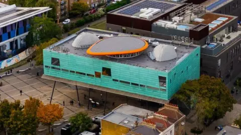 Getty Images An aerial view of Peckham Library, featuring a mix of residential, commercial, and cultural buildings. The library is a distinctive modern building with a bright turquoise-green exterior and a circular orange structure on its roof. The streets are lined with trees, and cars are visible parked along the roads.