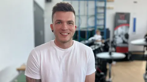 A man with brown hair is smiling at the camera. He has black stretchers in his earlobes and a tattoo sleeve on one arm, and is wearing a white t-shirt. Blurred in the background is a drumkit 