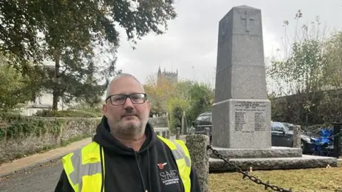 Andrew Stephenson wearing a black hooded jumper and a yellow hi-vis over the top. He has glasses and is smiling at the camera. Behind him on the right is the grey war memorial which has four blocks. At the top is cross at and names at the bottom. It is surrounded by grass and around the grass is concrete pillars with black chains.