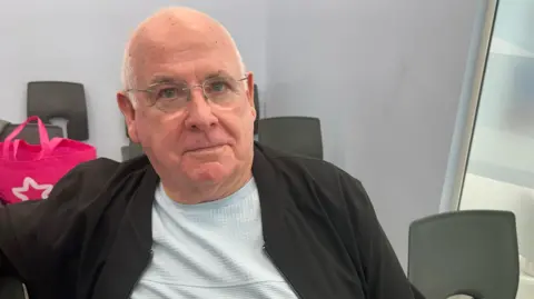 A man with a shaved head and glasses is looking at the camera. He is sitting amongst plastic chairs in a waiting room. There is a pink bag behind him.