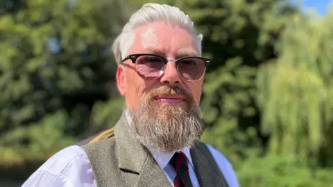 Emma Baugh/BBC A man with thick white hair swept back from his forehead and a beard. He is wearing glasses and a tweed jacket over a white shirt.