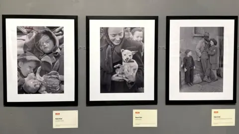 LEONARD MCCOMBE There framed photos on a wall featuring children holding a doll, a woman holding a small dog, and a man and a woman standing against a wall next to a child.