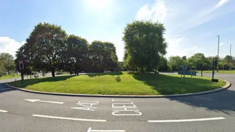 Google A screenshot from Google street view showing the A40 Benhall Roundabout in Cheltenham. It is a sunny day and the roundabout mound is covered in grass and several trees. There are no cars on the road at the time the picture was taken.