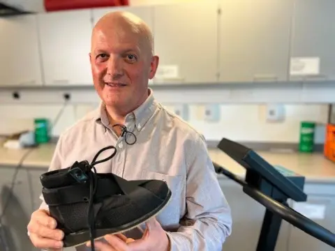 Prof Derek Santos, a bald man in a beige shirt stands in his lab. There is what looks like a running machine to his left.